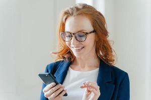 Intelligent positive female entrepreneur in elegant apparel, happy to recieve message on mobile phone, finds out about raising profits, has red hair, smiles happily, poses indoor in modern office photo