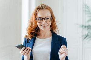 Photo of happy female leader with ginger hair, wears spectacles, updates app on modern cell phone, wears elegant clothes, poses in modern office, reads incoming message from business partner