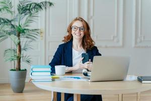 Horizontal shot of optimistic woman installs application on modern cell phone, checks notification, surrounded with paper documents, works on laptop computer, has red hair, dressed formally. photo