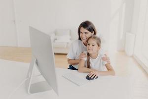 Indoor shot of happy lovely mother and daughter watch funny cartoon on computer, have pleasant smiles, enjoy togetherness, spend free time or weekend at home, spacious light empty living room photo