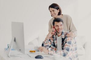Happy young man holds mobile phone, discusses issues, manages home accounts with wife, pose in coworking space, sit in front of computer, discuss family expenses. Couple collaborate together photo