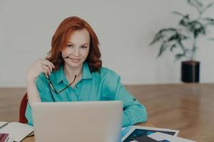 bella y feliz trabajadora independiente con cabello de jengibre, trabaja en línea, usa una computadora portátil, prepara un informe financiero en casa, usa una aplicación de videoconferencia, habla a distancia con sus colegas foto