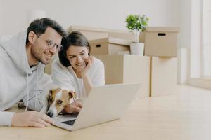 Glad young female and male discuss home repair projects, look attentively into laptop, their dog lies near, pose in living room on floor, dressed in casual clothes. People and new home concept photo