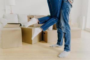 Cropped shot of caring man lifts his wife up, wears jeans and socks, just moved in new home, pose around cardboard boxes on floor, excited about moving. Unrecognizable couple relocate in new place photo