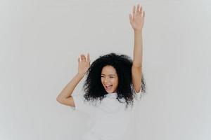 Joyful curly African American woman raises hands up, laughs from happiness, celebrates triumph, rejoices success, wears white t shirt, poses indoor, gestures actively, gets unexpected surprise photo