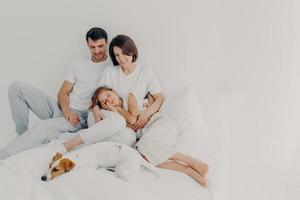 Photo of lovely family spend free time together, stay in bed for long time, had healthy sleep, pedigree dog lies near owners, enjoy comfort and relaxation, blank space on light background for promo