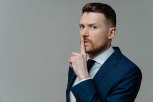 Serious bearded adult man makes shush gesture asks to be quiet tells confidential information presses index finger to lips wears formal clothes isolated over grey background with copy space. photo