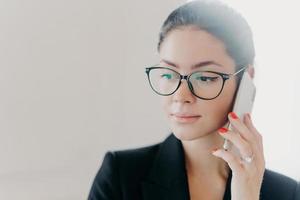 Headshot of serious brunette female manager with makeup wears trendy eyeglasses, formal clothes, has red manicure, calls business partner during remote job, poses indoor against white background photo