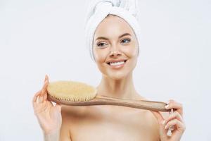 Young woman with healthy fresh skin, uses body brush, smiles gently, wears bath towel on head, poses topless, isolated over white background. Spa accessories. Women, body care, hygiene concept photo