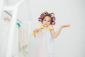 Hesitant girl with clueless expression, doesnt know what to wear, holds hangers with dress, has curlers on head, isolated over white background. Cute child with pleasant appearance prepares for party photo