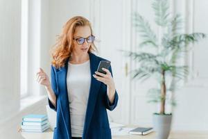 Waist up shot of successful businesswoman uses application on cell phone, reads recieved email from boss or colleague, dressed in formal clothes, poses over office interior, finds out financial news photo