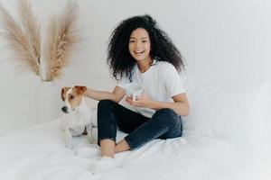 Happy relaxed lovely young woman with Afro hairstyle, petting dog, sit together on bed with white bedclothes, drink hot beverage, dressed in casual wear, expresses positive emotions. People and rest photo
