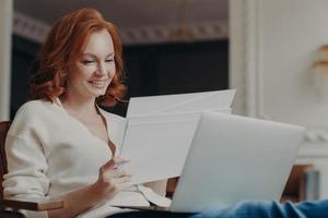 Horizontal view of pleased adult woman entrepreneur works from home, works remotely from home, looks through paper documents, uses laptop computer, wears casual jumper, poses in own cabinet. photo
