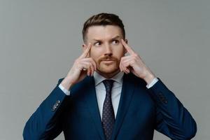 toma interior de un hombre de negocios barbudo, motivado y pensativo, que mantiene los dedos en las sienes, mira hacia otro lado, se prepara para la conferencia vestido con ropa formal aislada sobre fondo gris, tiene un duro día de trabajo foto