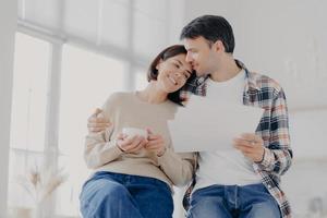 Indoor shot of relaxed beautiful woman with coffee cup, receives hug from husband who holds papers, make financial report together, own business company. Family, finances and budget concept. photo
