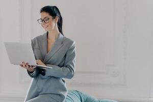 Happy female entrepreneur types on laptop computer, works in modern workspace in business center, dressed in formal outfit, surfes internet, watches training webinar, chats online with partners photo