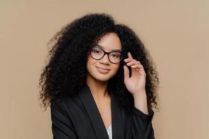 Close up portrait of calm satisfied Afro American woman with bushy curly hairstyle, keeps hand raised, has healthy skin, wears spectacles, black elegant outfit isolated on brown wall. Face expressions photo