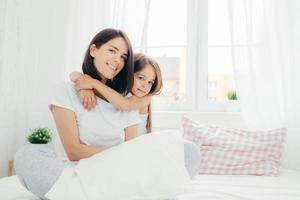 People, family and bedding concept. Cheerful young smiling mother and her daughter embrace each other, have positive expression, wear pyjamas, sit on comfortable bed against window background.