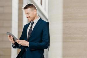 Prosperous businessman in formal clothes reads notification on digital tablet, installs new application, connected to wireless internet, poses indoor. People, business and modern technology concept photo