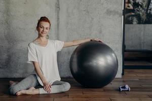 Happy young fitness woman relaxing after pilates workout while sitting with large exercise ball on floor photo