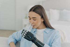 Girl is using futuristic arm prosthesis holding glass of water. Healthy lifestyle after amputation. photo