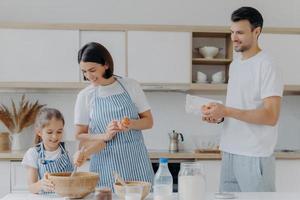 Happy parents cook together with daughter, pose at modern home kitchen give eggs to add to dough, small kid whisks ingredients in bowl have glad expressions. Food and family concept. Cooking breakfast photo