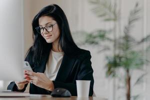 Satisfied business lady uses smartphone, poses at work place in front of computer screen, cup of tasty beverage, sends messages during break, poses at contemporary office. Technology and work concept photo