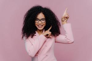 Half length shot of cheerful Afro American woman with dark bushy hairstyle, wears violet suit, points above with both index fingers, demonstrates way to something, invites you going upstairs photo