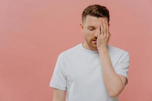 Horizontal shot of serious tired man makes face palm feels bored spending time alone covers eye with hand dressed in casual white t shirt isolated over pink background blank space on right side photo