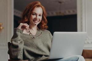 Professional positive redhead young European woman works on freelance, concentrated on remote job, prepares publication for web page, sits in front of opended laptop computer, smiles gently. photo