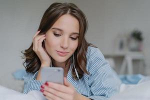 Indoor shot of pretty dark haired woman enjoys music in playlist, lies on comfortabled bed at home, has day off, watches films online, connected to wireless internet, enjoys cozy atmosphere. photo