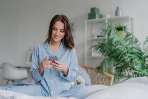 Indoor shot of happy dark haired woman in casual domestic costume, enjoys rest time, poses in cozy bedroom, enjoys music with modern devices, domestic interior in background. People, lifestyle concept photo