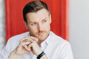 Pensive male director keeps hands presssed together, looks thoughtfully aside, being deep in thoughts before important meeting, poses indoor against red and white wall with copy space for your text photo