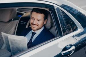Handsome young banker in smart formal tuxedo suit reads newspaper in luxury auto