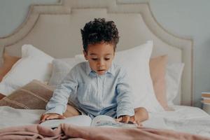 Intrigued afro american preschool child looking at colorful pictures inside of book while sitting on big bed photo