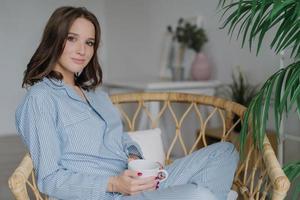 Sideways shot of pretty woman sits crossed legs in wicker chair, dressed in pyjamas, drinks hot coffee and latte, poses against cozy interior indoor, has weekend. People and relaxation concept photo