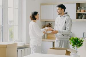 Image of delighted family couple unpack utensils with carton boxes, move in new home, pose against kitchen interior, looks happily at each other, busy unpacking different home stuff. Householding photo