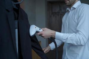 Businessman preparing clothes for meeting with partner, steaming suit with portable steamer photo