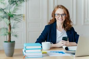 Photo of red haired smiling young woman installs app on mobile phone, checks newsfeed, works on laptop computer remotely, poses in coworking space with cup of drink, books and paper documents