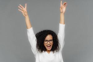 Happy pleased young woman raises hands up, being in high spirit, dances over grey background, celebrates someting, has Afro haircut, wears white jumper, transparent glasses for vision correction photo