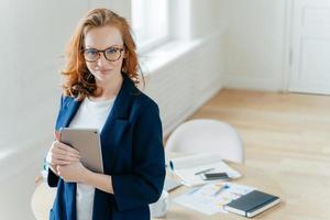 Shot of beautiful red haired young woman checks notification on touch pad, happy to recieve income message, looks confidently at camera, workplace with paper documents and notepad in background. photo