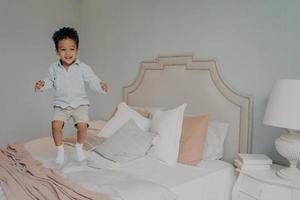 Happy carefree mixed race ethnicity kid boy in casual clothes having fun and jumping on bed photo