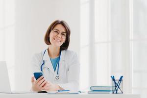 Horizontal view of smiling professional doctor works in clinic, poses at modern hospital office with electronic gadgets, sends text messages on cellphone being at work. Health care, technology concept photo