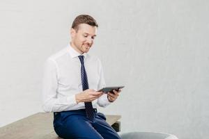 Positive male financier wears white shirt, tie, sits on table, installs new application on touch pad, connected to wireless internet. Successful businessman reads notification on modern device photo
