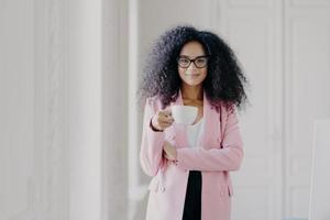 Shot of attractive woman with Afro hairstyle, drinks aromatic beverage, wears glasses, formal wear, poses in office over white background, copy space for your promotional content. People and business photo