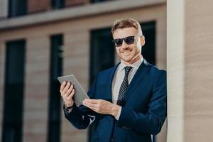 Portrait of happy male entrepreneur sends text message to colleague via touch pad, connected to wireless internet, updates software, wears trendy sunglasses and black suit. People and technology photo