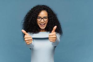 Happy lovely woman with bushy Afro hair gives thumb up, approves nice idea, wears fashionable sweater, isolated over blue background. Its fine, I agree. Body language concept. People and like concept photo