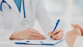 Unrecognizable woman doctor writes down in clipboard, makes prescription for patient, records data for analysis, wears white gown. Close up, focus on hands. Medicine, insurance, health care concept photo