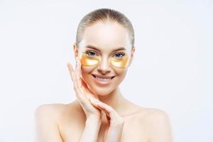 Studio shot of attractive European woman touches face gently, smiles tenderly, applies golden hydrogel patches under eyes, isolated on white background. Eye mask treatment. Anti age procedure photo