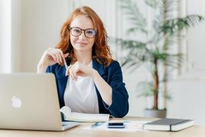 Positive red haired young female entrepreneur watches business webinar, writes email, makes notes in notepad, wears optical glasses, dressed in formal clothes, works online, involved in banking sphere photo